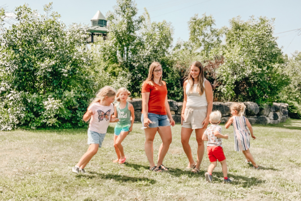 Meghan Magritz and Allie Wedl stand on a lawn with four of their kids running around them.