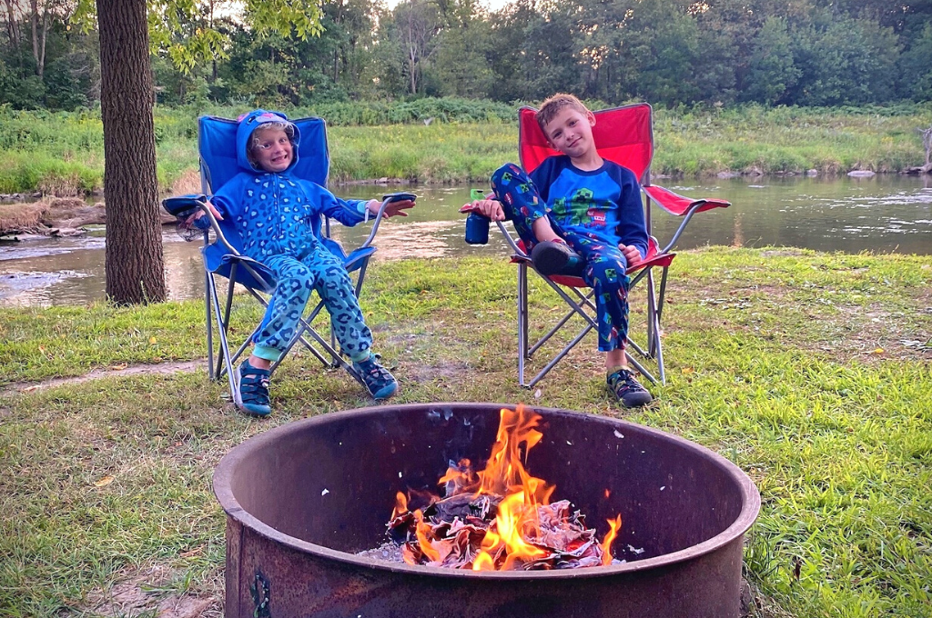 Two kids sit in camp chairs by a campfire.